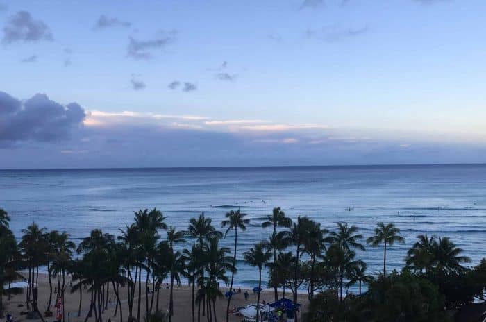 The image shows Waikiki Beach right after sunrise. There are palm trees on the beach in front of the ocean and clouds in the sky.