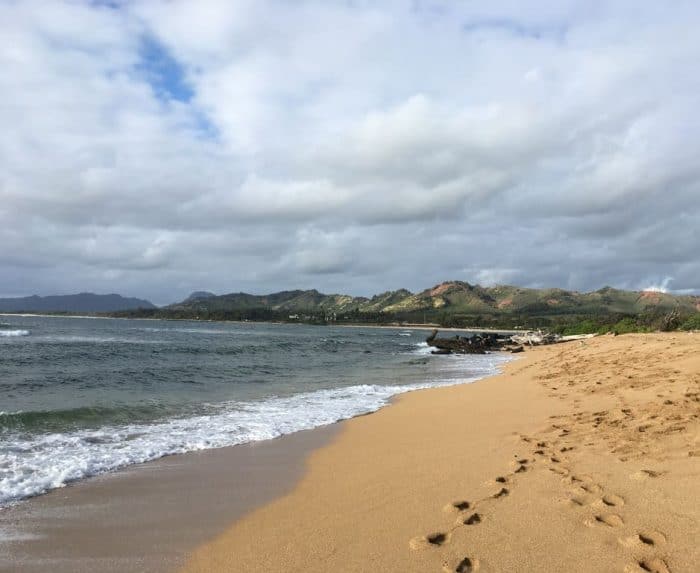 A sandy beach on Kauai with waves receding from the shore, a cloud-filled sky, and green and red dirt hills in the background.