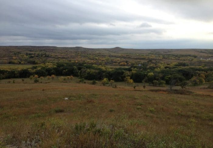 The image shows a cloudy sky over a prairie landscape in the fall. The trees are changing colors and the grass is a mix of green, red, and brown.